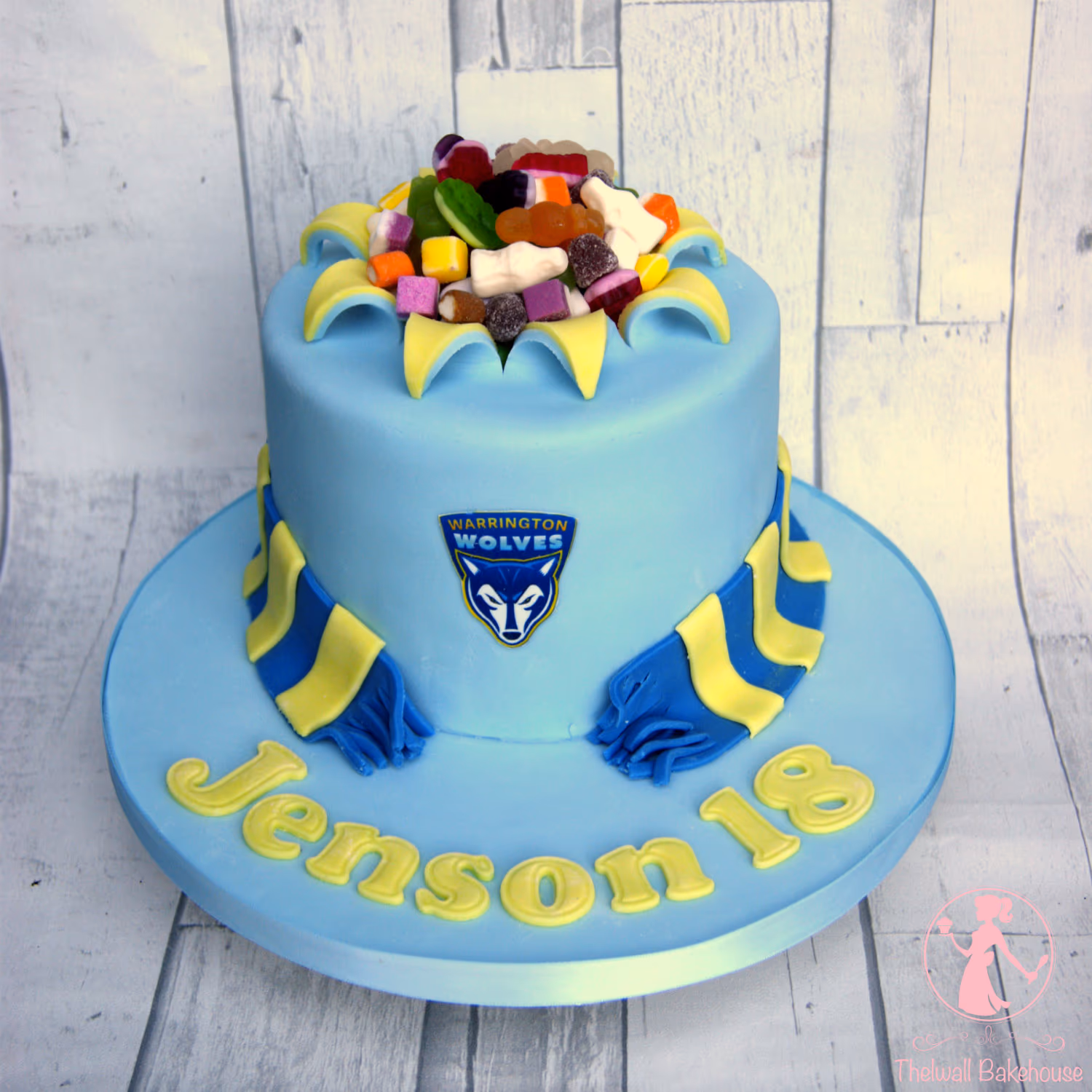 A light blue round birthday cake for Jenson's 18th birthday, decorated with the Warrington Wolves logo, a blue and yellow striped scarf, and topped with a variety of colorful sweets.
