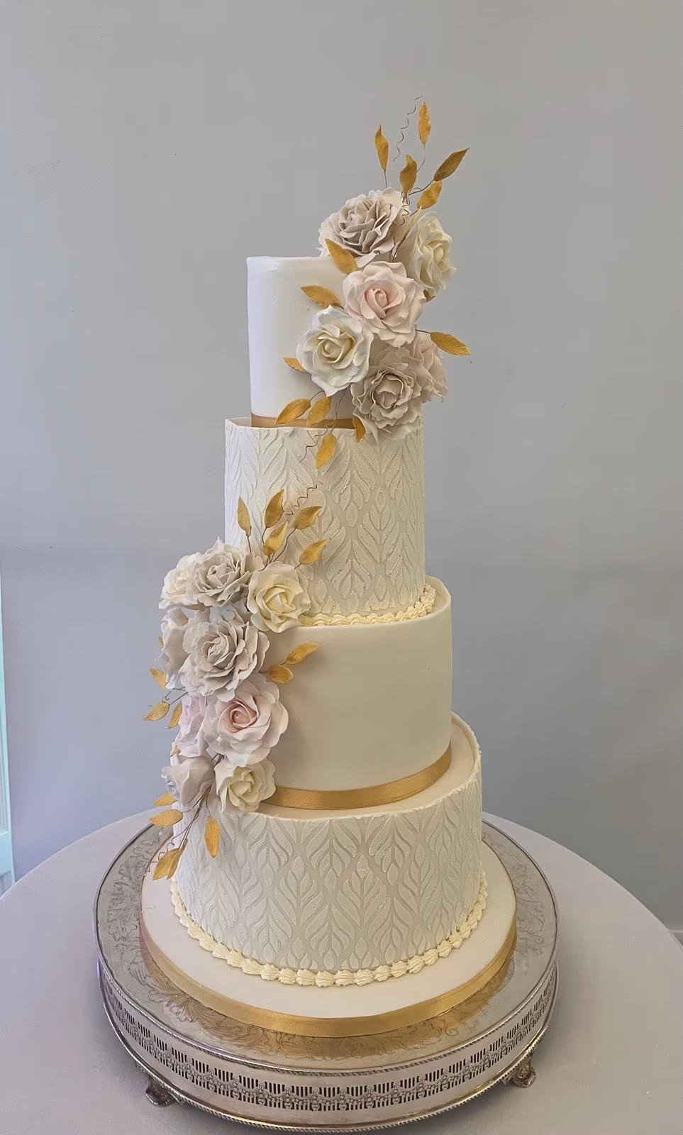 Elegant four-tier wedding cake with alternating smooth and patterned cream fondant tiers, decorated with gold ribbons and a cascading arrangement of pastel sugar roses and gold leaves.