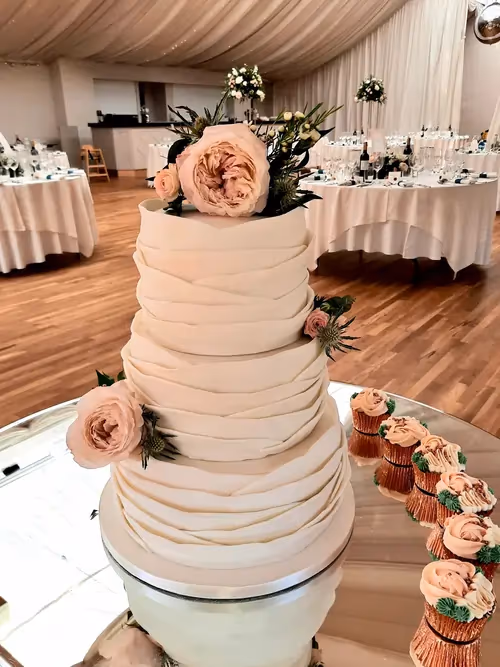 A four-tier wedding cake with white draped fondant, decorated with light pink roses and green thistles, displayed on a mirrored stand next to matching cupcakes in a wedding reception venue.