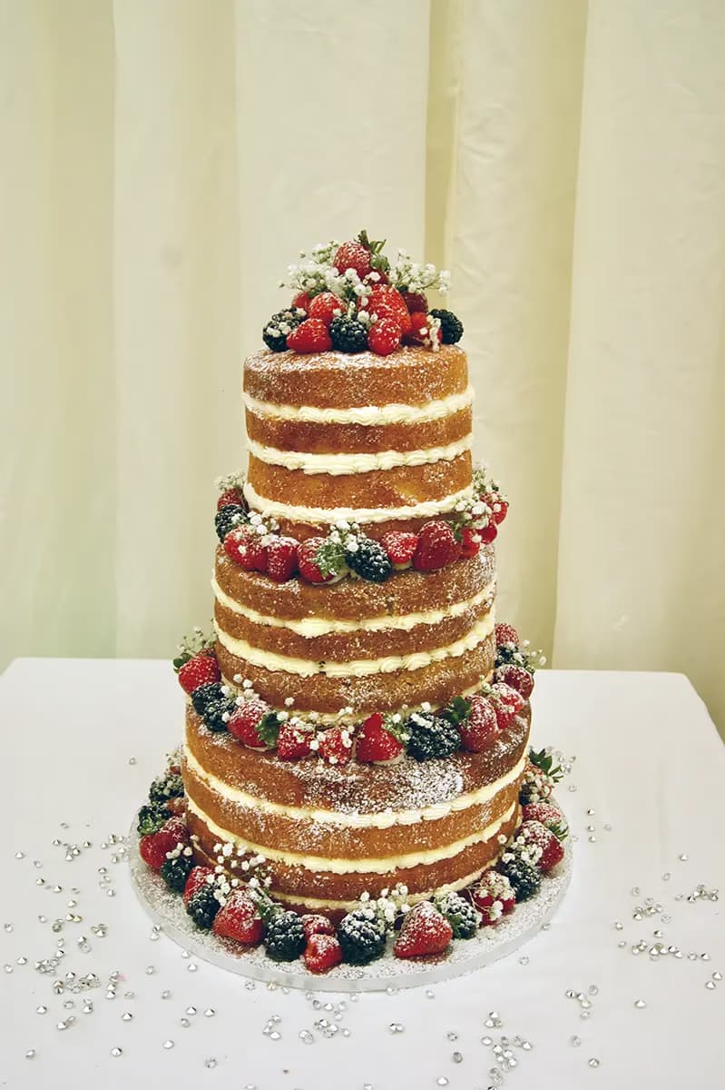 A four-tier naked wedding cake featuring visible sponge layers and white cream filling, generously decorated with fresh mixed berries including strawberries, raspberries, and blackberries, interspersed with delicate white baby's breath flowers and dusted with powdered sugar, set on a white table with silver confetti.