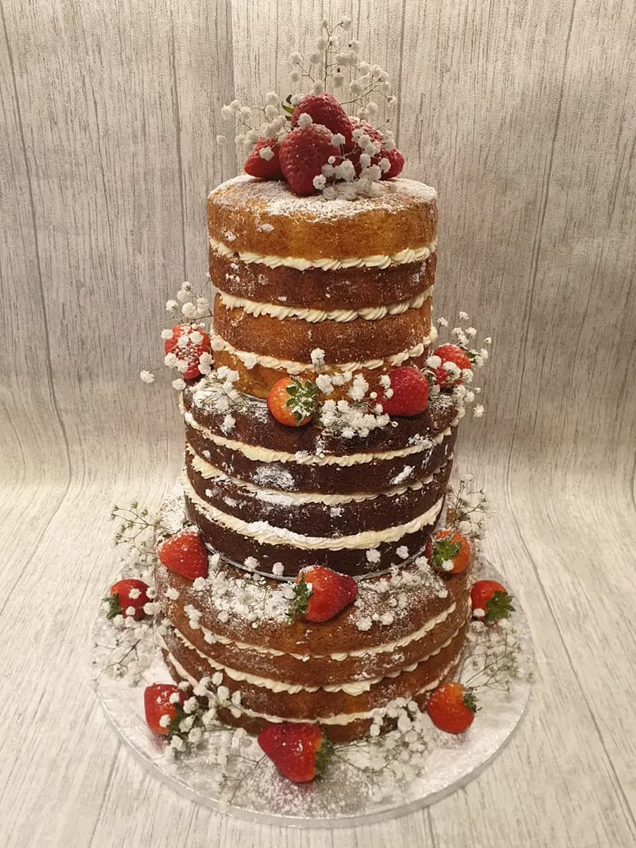 A four-tier naked wedding cake featuring visible sponge and cream layers, generously decorated with fresh strawberries, delicate white baby's breath flowers, and a dusting of powdered sugar.