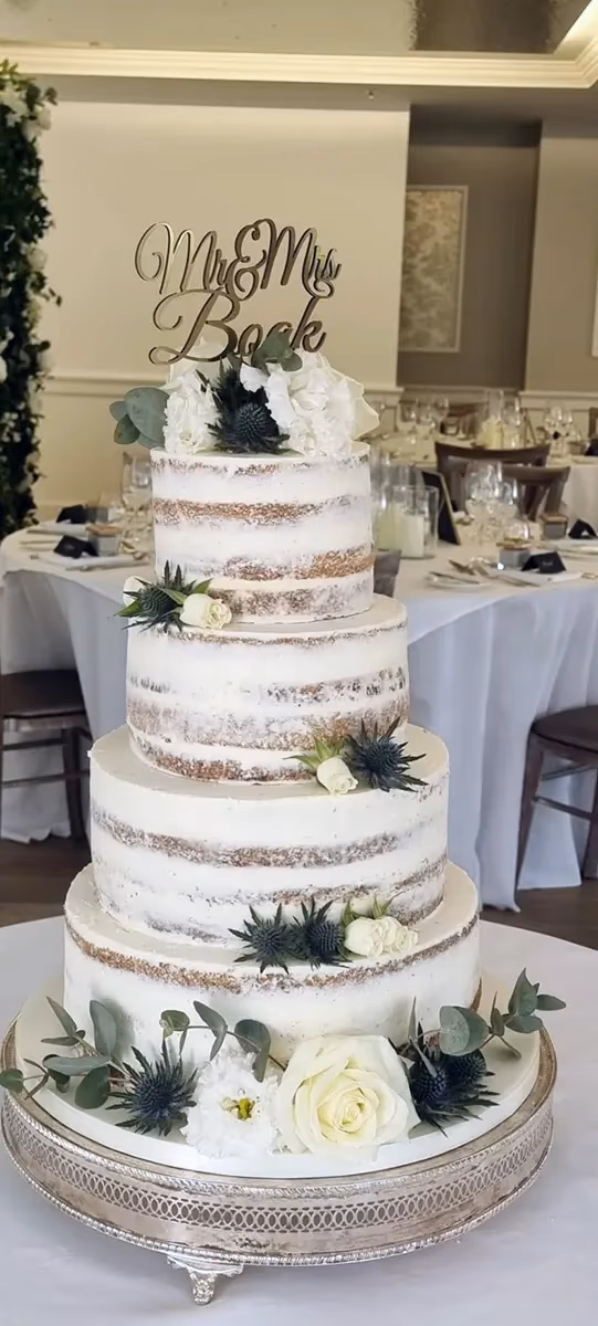 A four-tier naked wedding cake decorated with white flowers, thistles, and greenery, topped with a 'Mr & Mrs Book' cake topper.