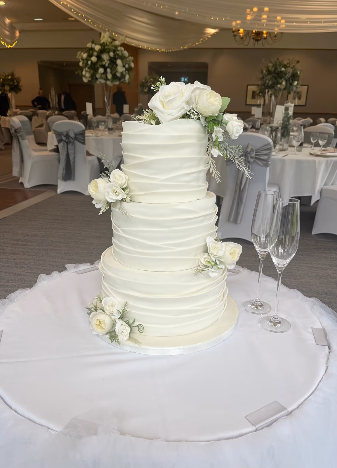 A four-tier white wedding cake with a draped fondant design, adorned with white and cream flowers and silver-green foliage, displayed on a white table with two champagne flutes in a wedding reception venue.