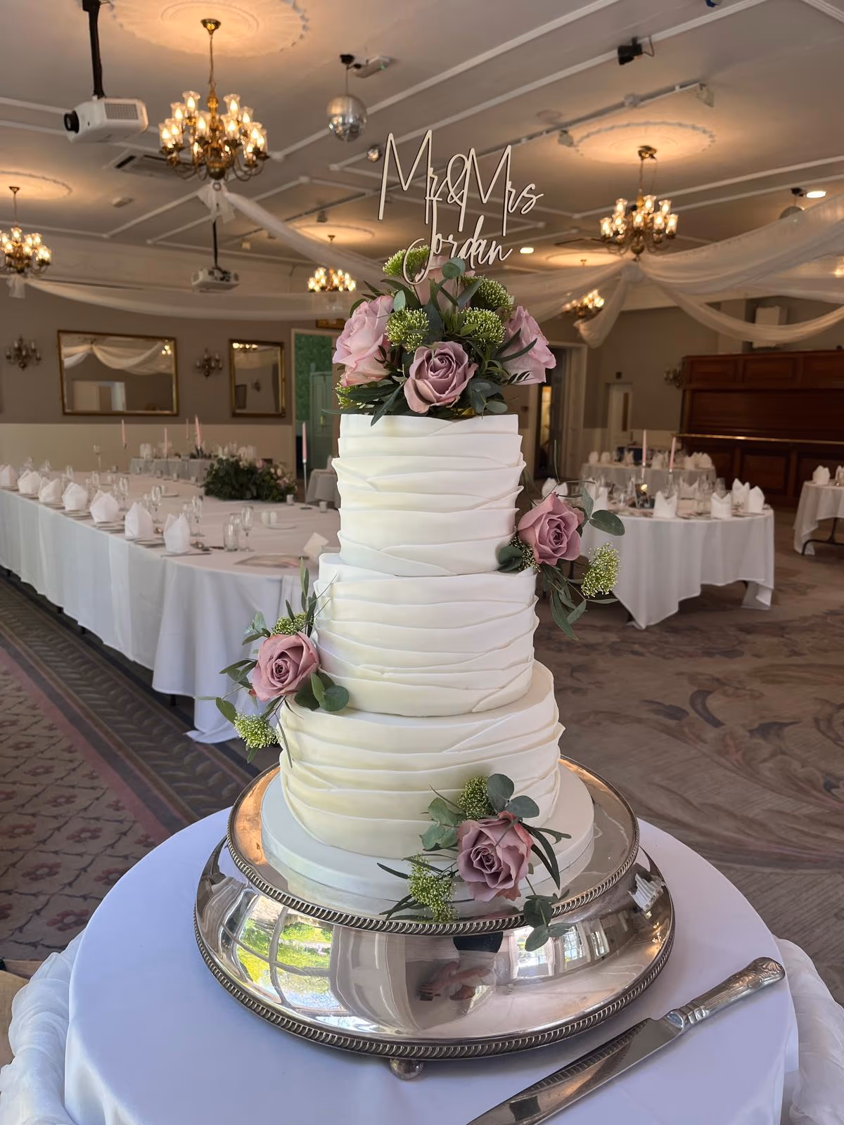 A four-tier white wedding cake with ruffled fondant, decorated with pink and mauve roses and green foliage, topped with a 'Mr & Mrs Jordan' sign, set on a silver stand in a wedding reception venue with tables and chandeliers in the background.