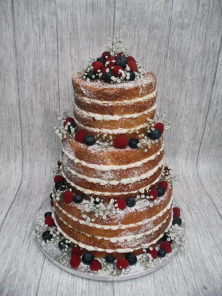 A three-tier naked wedding cake is decorated with fresh raspberries, blueberries, baby's breath flowers, and a dusting of powdered sugar.