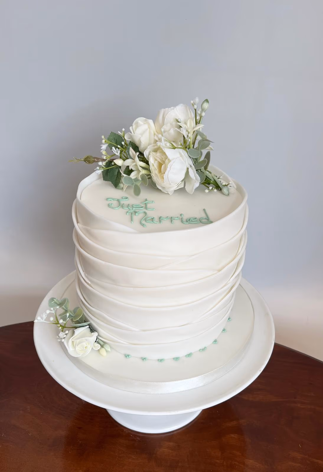 A single-tier white wedding cake with a draped fondant texture, topped with a bouquet of white roses and green foliage. The words 'Just Married' are written in light green script on the top of the cake. The cake sits on a white stand, with a small white rose and green leaves on the base, accompanied by a line of light green heart-shaped dots.
