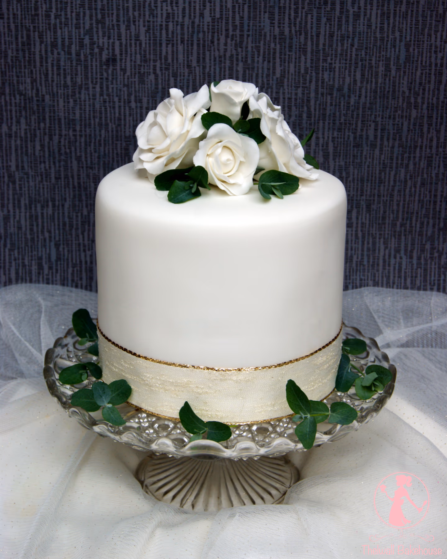 A single-tier white fondant wedding cake adorned with white sugar roses and eucalyptus leaves on top, a cream and gold ribbon around its base, sitting on a clear glass cake stand.