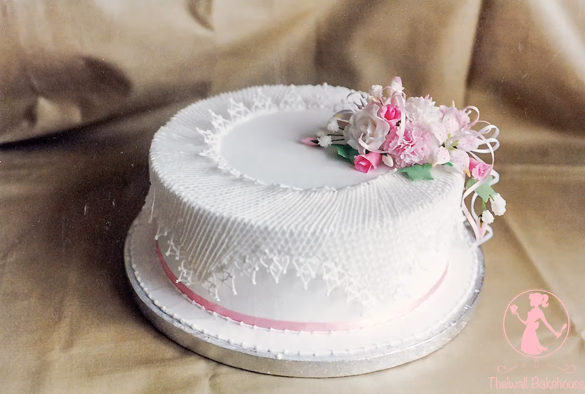 A single-tier white wedding cake featuring intricate white lace piping on the sides and top, adorned with a cluster of pink and white sugar flowers and a pink ribbon around its base.