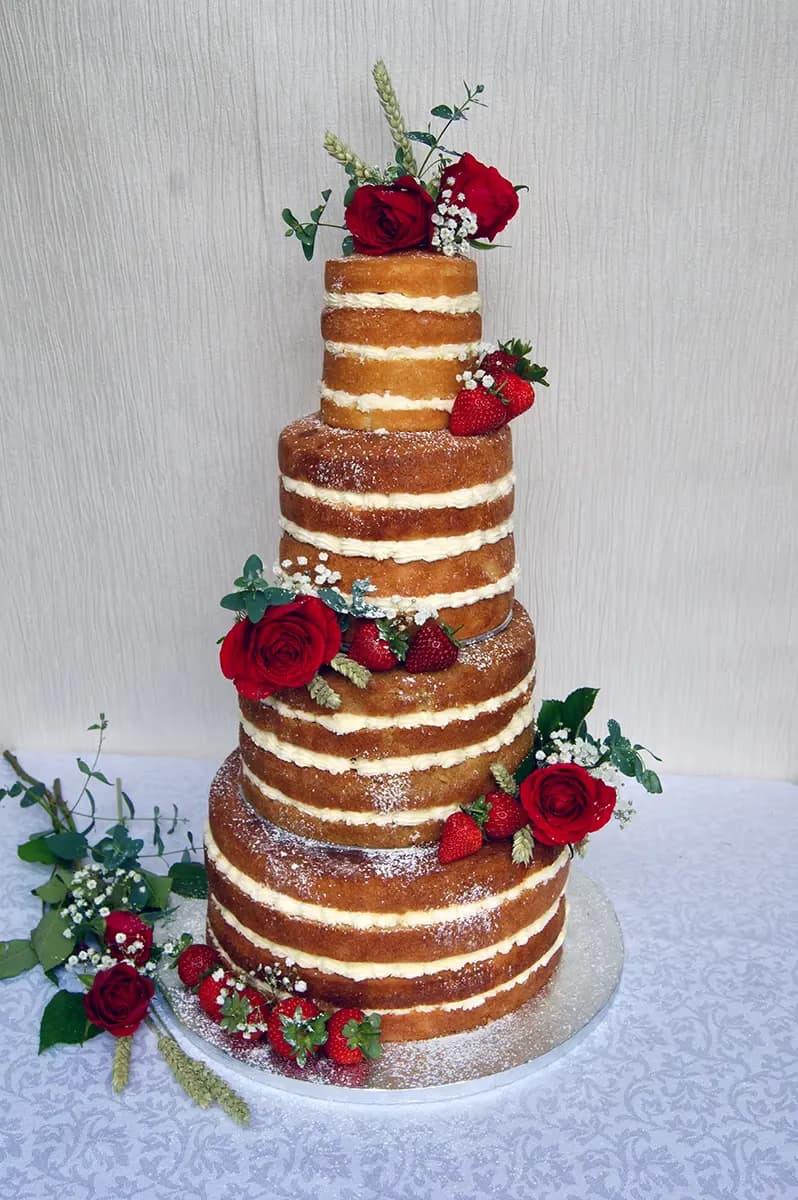 A six-tier naked wedding cake decorated with red roses, fresh strawberries, greenery, and wheat stalks, lightly dusted with powdered sugar.