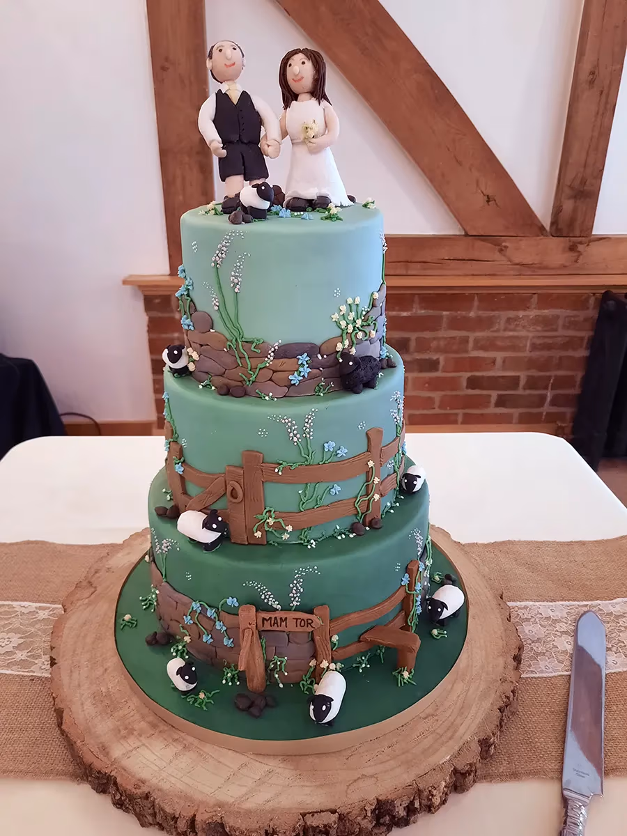 A three-tier wedding cake with green fondant, decorated with fields, stone walls, wooden fences, blue and white flowers, and several sheep figurines, topped with bride and groom figures, and featuring a 'Mam Tor' sign on the bottom tier, displayed on a wooden tree slice.