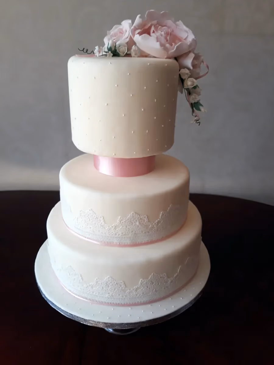 A three-tier wedding cake with cream fondant, featuring pink sugar flowers cascading down the top tier, white lace patterns on the lower tiers, and delicate pink ribbon accents.