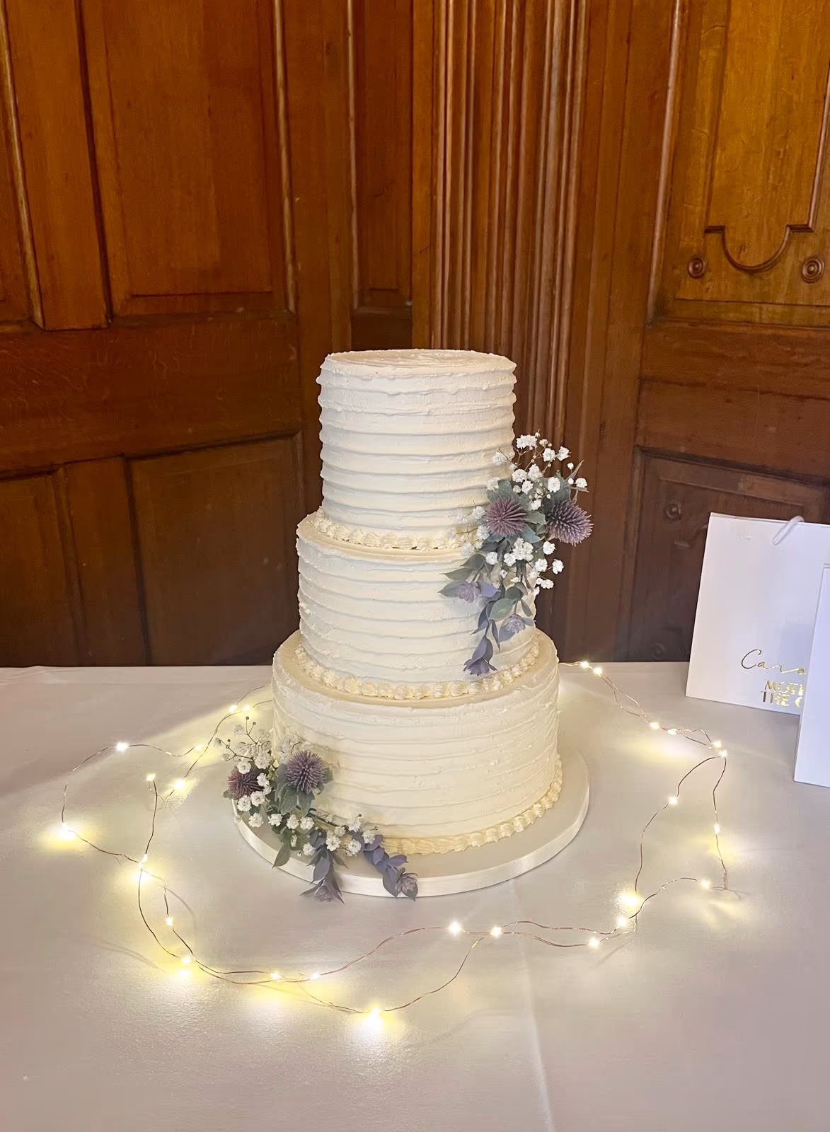 A three-tier cream-colored wedding cake with textured buttercream frosting, decorated with purple thistles, small white flowers, and green foliage cascading down the side. Fairy lights are draped around the base of the cake on a white tablecloth, with a wooden paneled background.