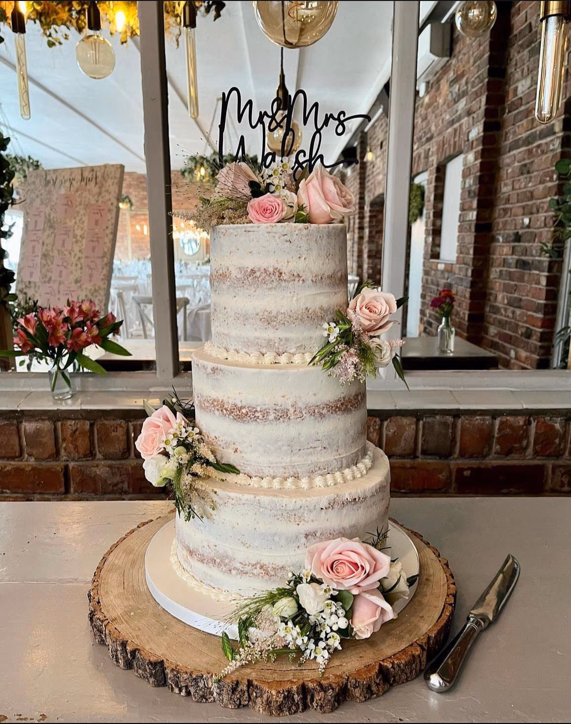 A three-tier naked wedding cake decorated with blush pink roses, small white flowers, and greenery, topped with a 'Mr & Mrs Walsh' cake topper, sitting on a rustic wooden log slice.