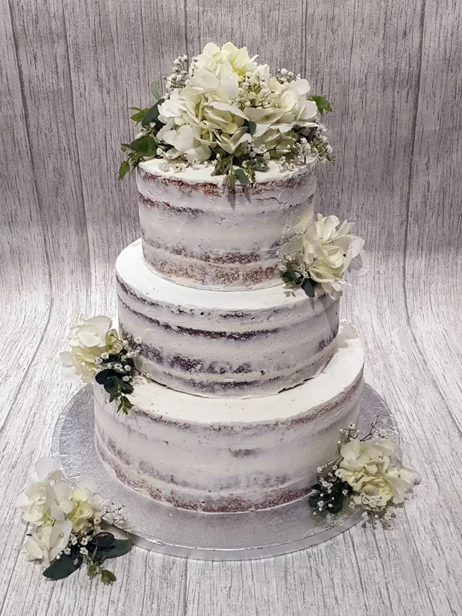 Three-tier naked wedding cake decorated with white hydrangeas, baby's breath, and green foliage.