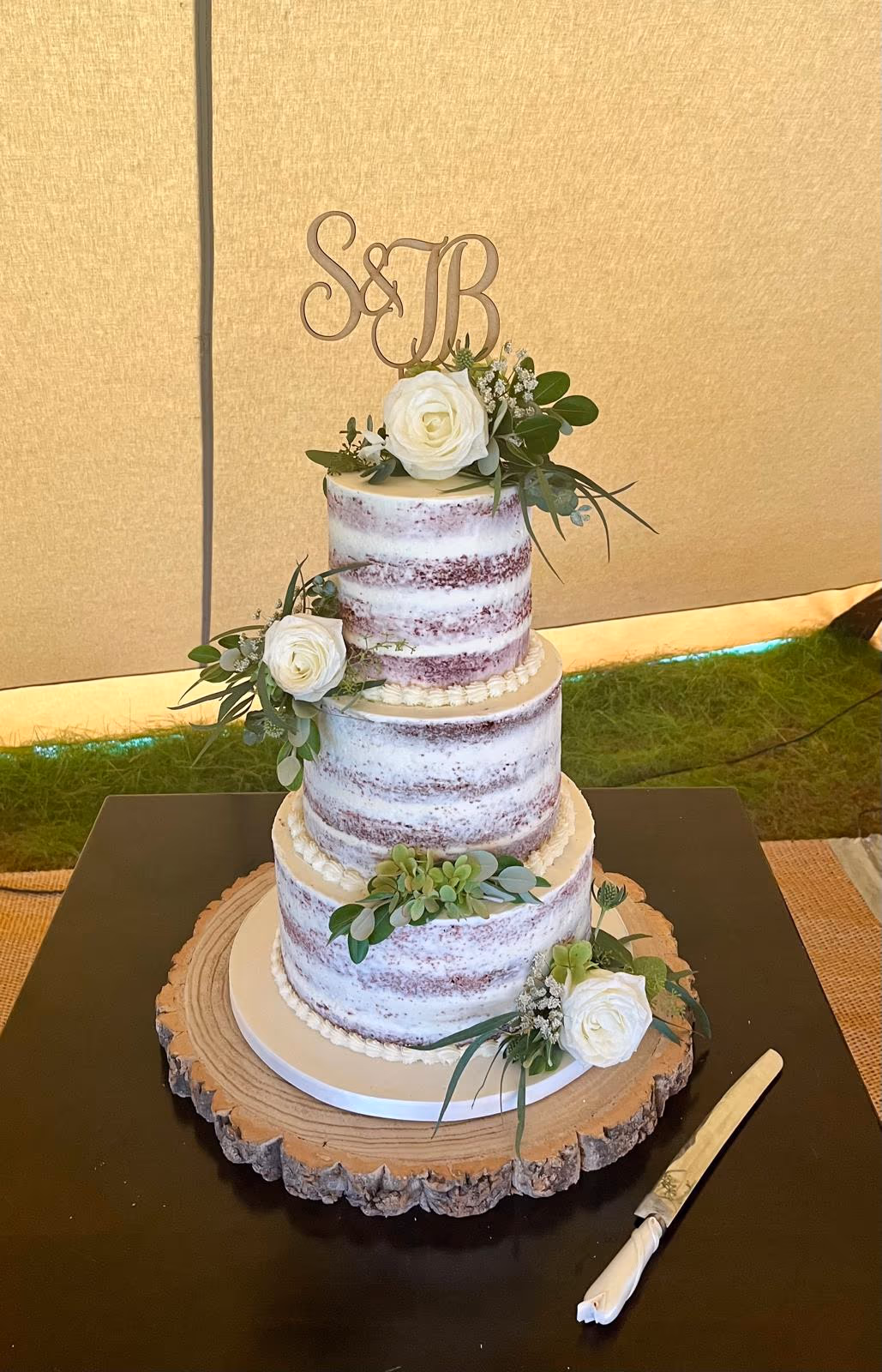 Three-tier naked wedding cake decorated with white roses, assorted greenery, and a wooden 'S & B' initial topper, presented on a rustic wooden cake stand.