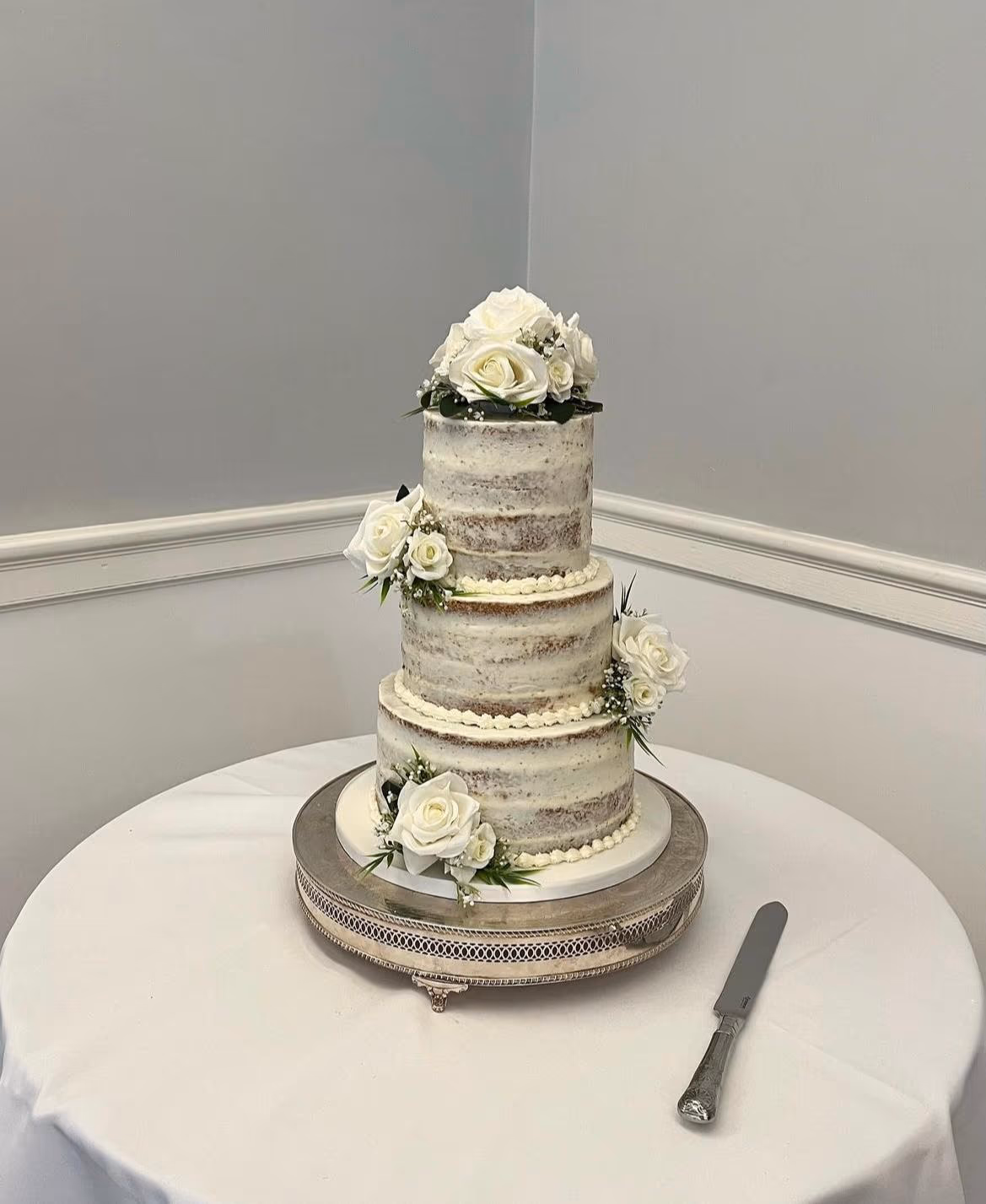 Three-tier naked wedding cake decorated with white roses and green foliage, presented on a silver cake stand.