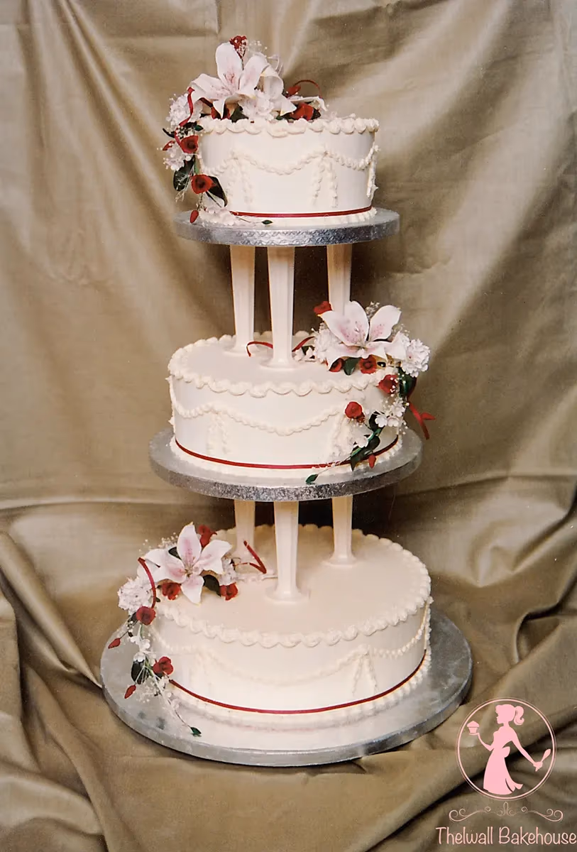 A three-tier white wedding cake with pillars, adorned with pink lilies, red roses, and white accent flowers, red ribbons, and delicate piped swags.