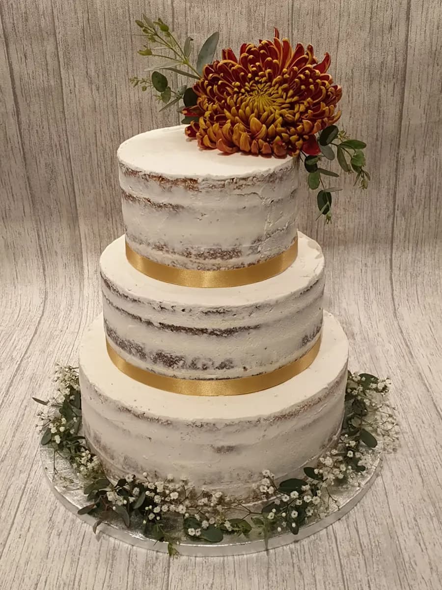 A three-tier semi-naked wedding cake with white frosting, gold ribbons, a large red and orange chrysanthemum, and green foliage, with baby's breath around the base.