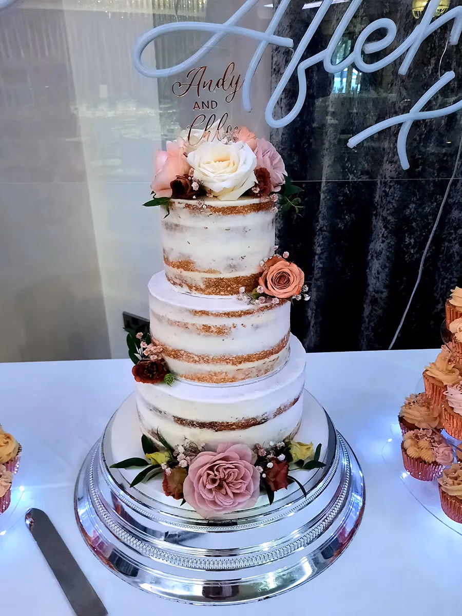 A three-tier semi-naked wedding cake with visible cake layers through a thin white buttercream, decorated with pink, white, and burgundy roses, greenery, and small white filler flowers. A gold cake topper reads 'Andy and Chloe'. The cake sits on a silver tiered stand with string lights, and in the background, a neon sign partially reads 'After'.