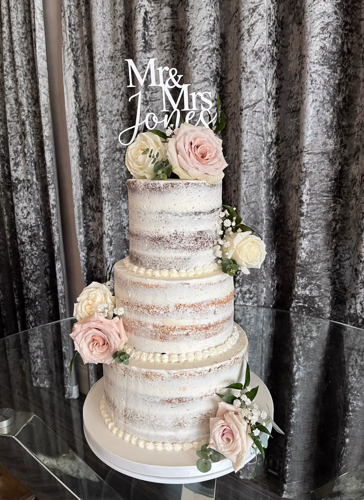 A three-tier semi-naked wedding cake decorated with white and light pink roses, greenery, baby's breath, and a 'Mr & Mrs Jones' cake topper, displayed on a glass table.