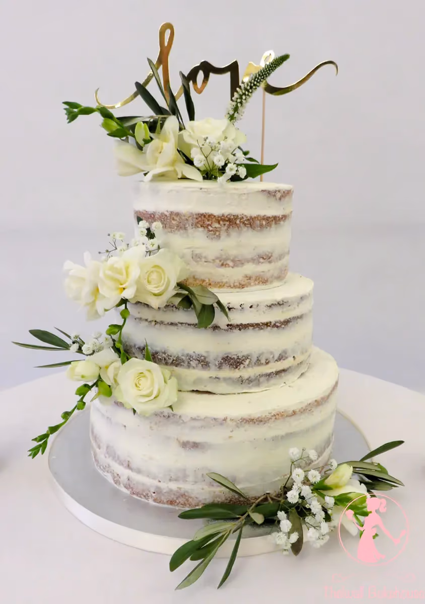 A three-tier semi-naked wedding cake adorned with white roses, baby's breath, green foliage, and a gold 'love' cake topper.