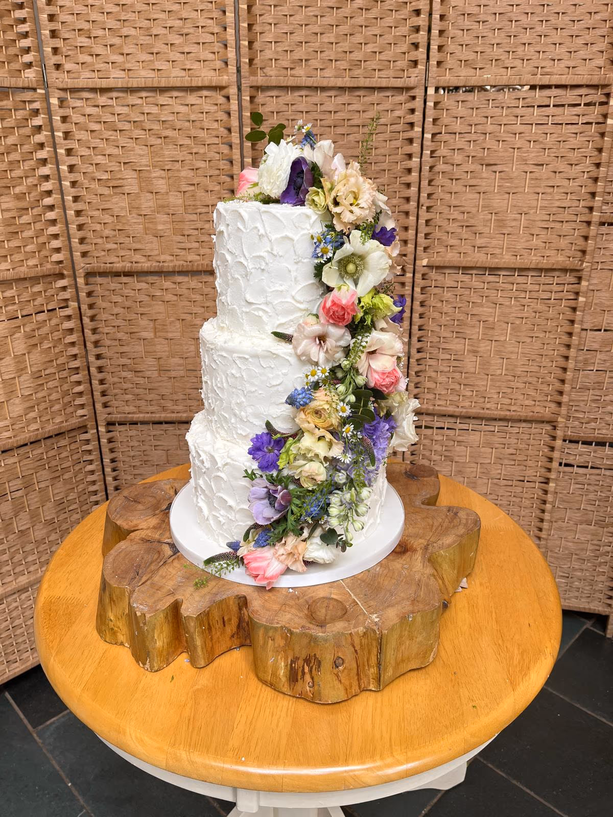 Three-tier white wedding cake with a textured buttercream finish, adorned with a vibrant cascade of fresh multi-colored flowers, presented on a rustic wooden log stand.
