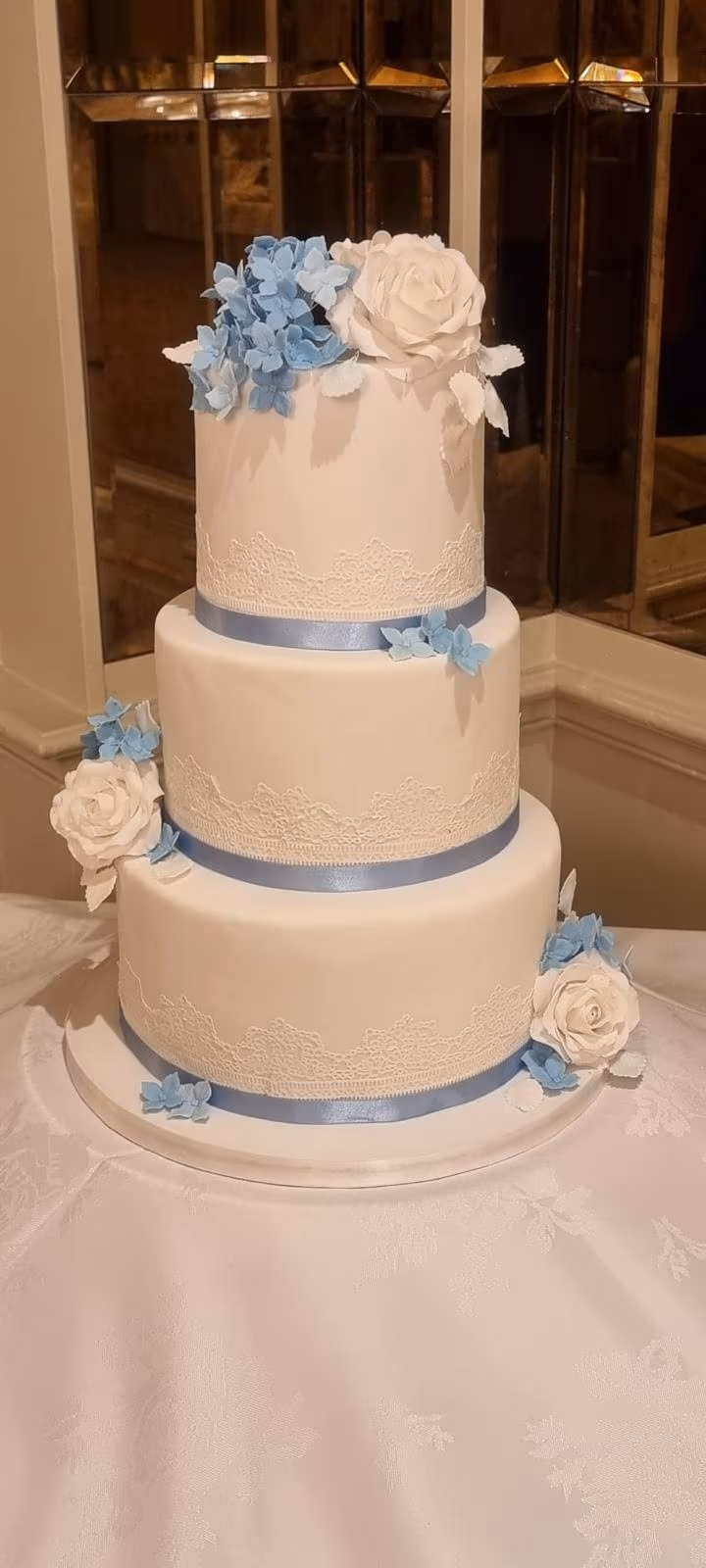 A three-tier white fondant wedding cake decorated with light blue satin ribbons, white lace detailing, blue hydrangea flowers, and white sugar roses, displayed on a white tablecloth.