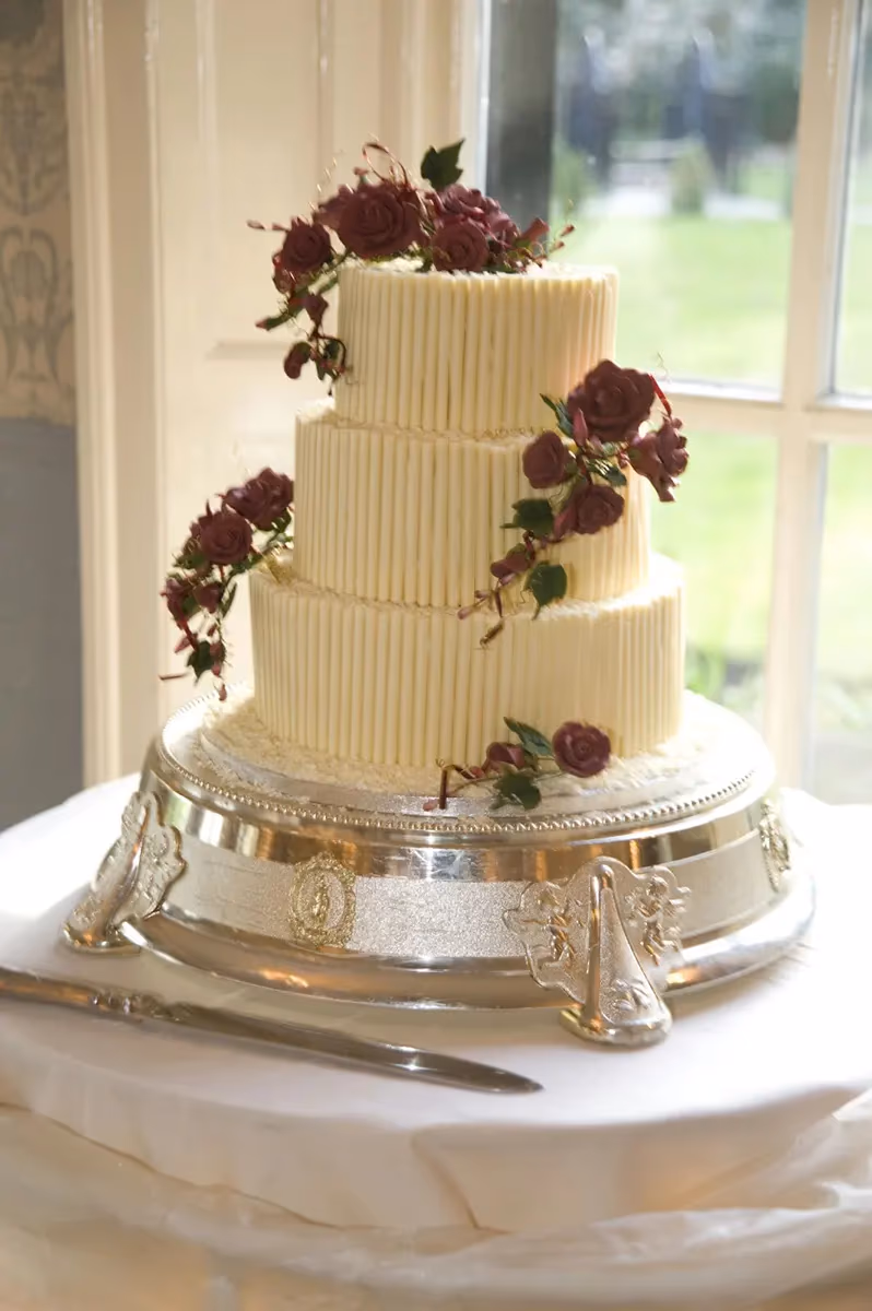 A three-tier wedding cake decorated with vertical white chocolate sticks and cascading deep red sugar roses, presented on an ornate silver cake stand with a cake knife nearby.