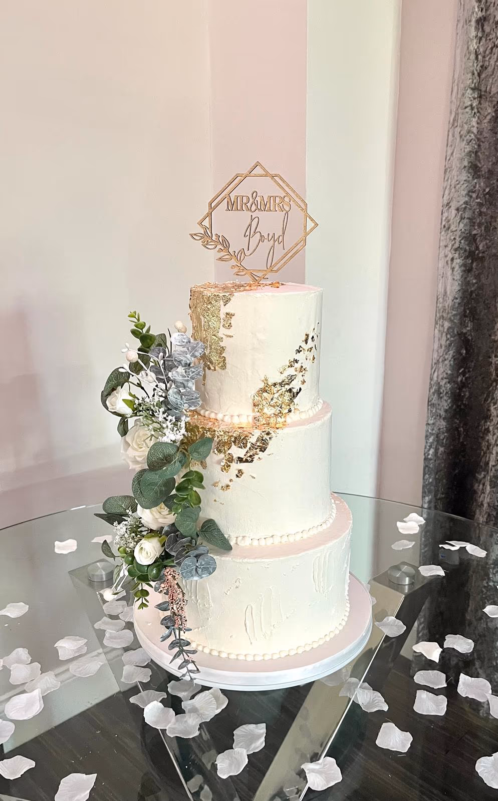A three-tier wedding cake with white frosting and gold leaf accents, decorated with a cascading floral arrangement of white roses, green eucalyptus, and blue-grey foliage, and topped with a gold 'MR & MRS Boyd' topper.
