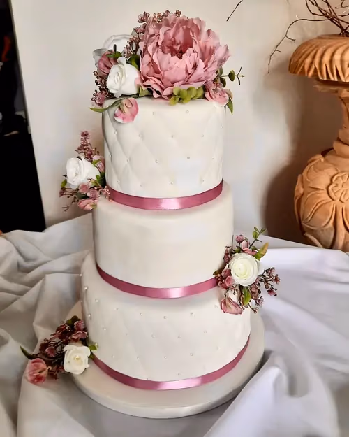 A three-tier white wedding cake featuring large pink and smaller white sugar flowers, pink satin ribbons, and a quilted pattern with edible pearls on the top and bottom tiers.