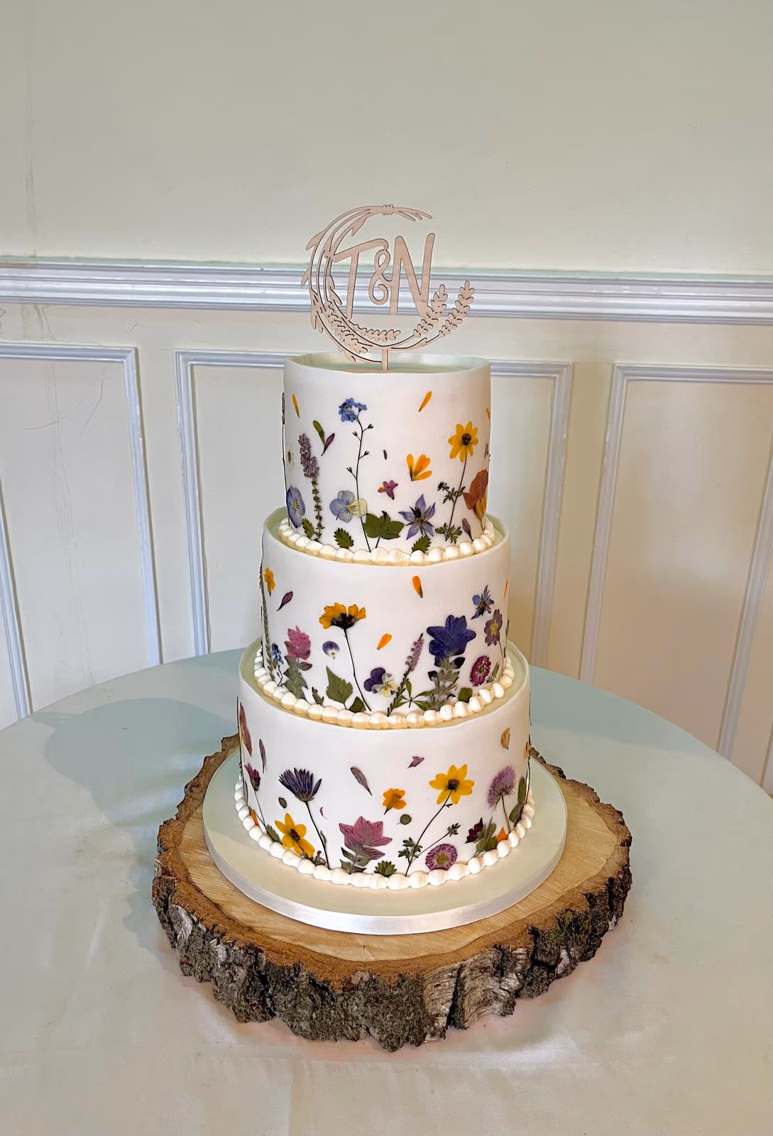 A three-tier white wedding cake adorned with colorful pressed wildflowers and a wooden 'T & N' initial cake topper, displayed on a rustic tree stump stand.