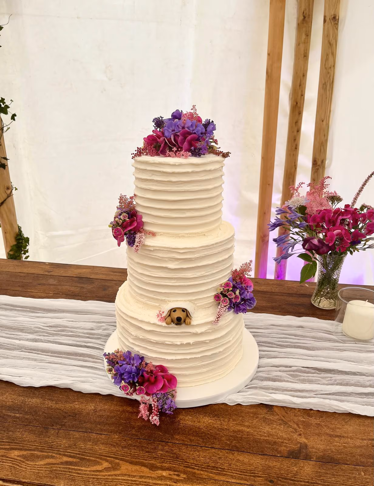 A three-tier white wedding cake with ribbed buttercream frosting, adorned with vibrant purple and pink floral clusters, and featuring a small brown dog peeking from a cutout on the bottom tier, set on a rustic wooden table.