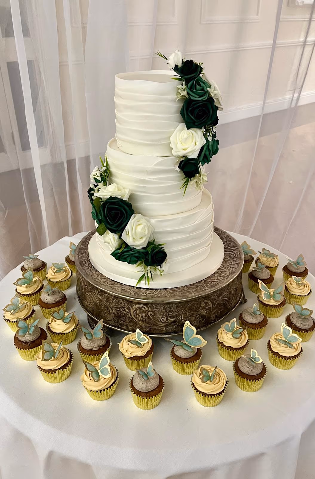 A three-tier white wedding cake with horizontal ruffled frosting, decorated with a cascade of dark green and white roses and greenery, surrounded by cupcakes with light blue butterfly toppers.