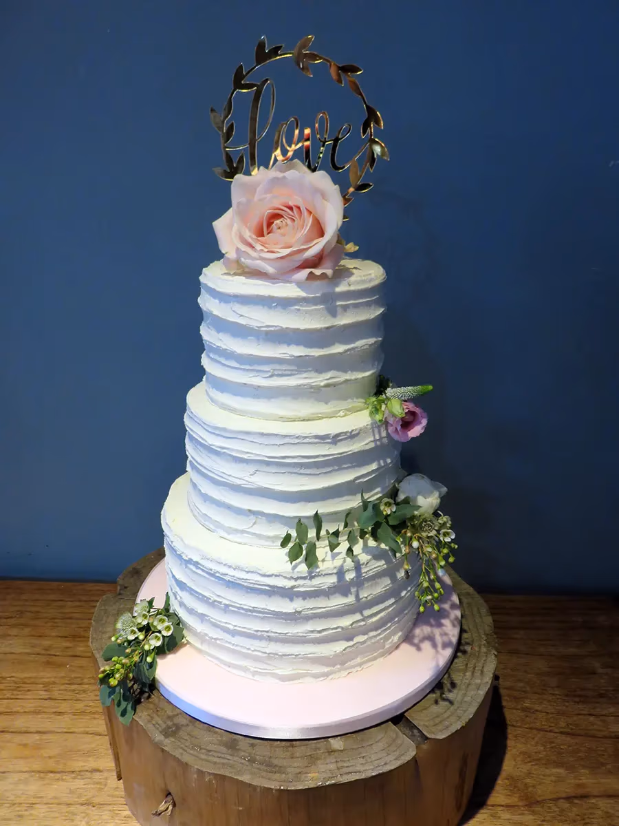 A three-tier wedding cake with white textured buttercream, topped with a pink rose and a gold 'love' topper, and decorated with green foliage and small blossoms.