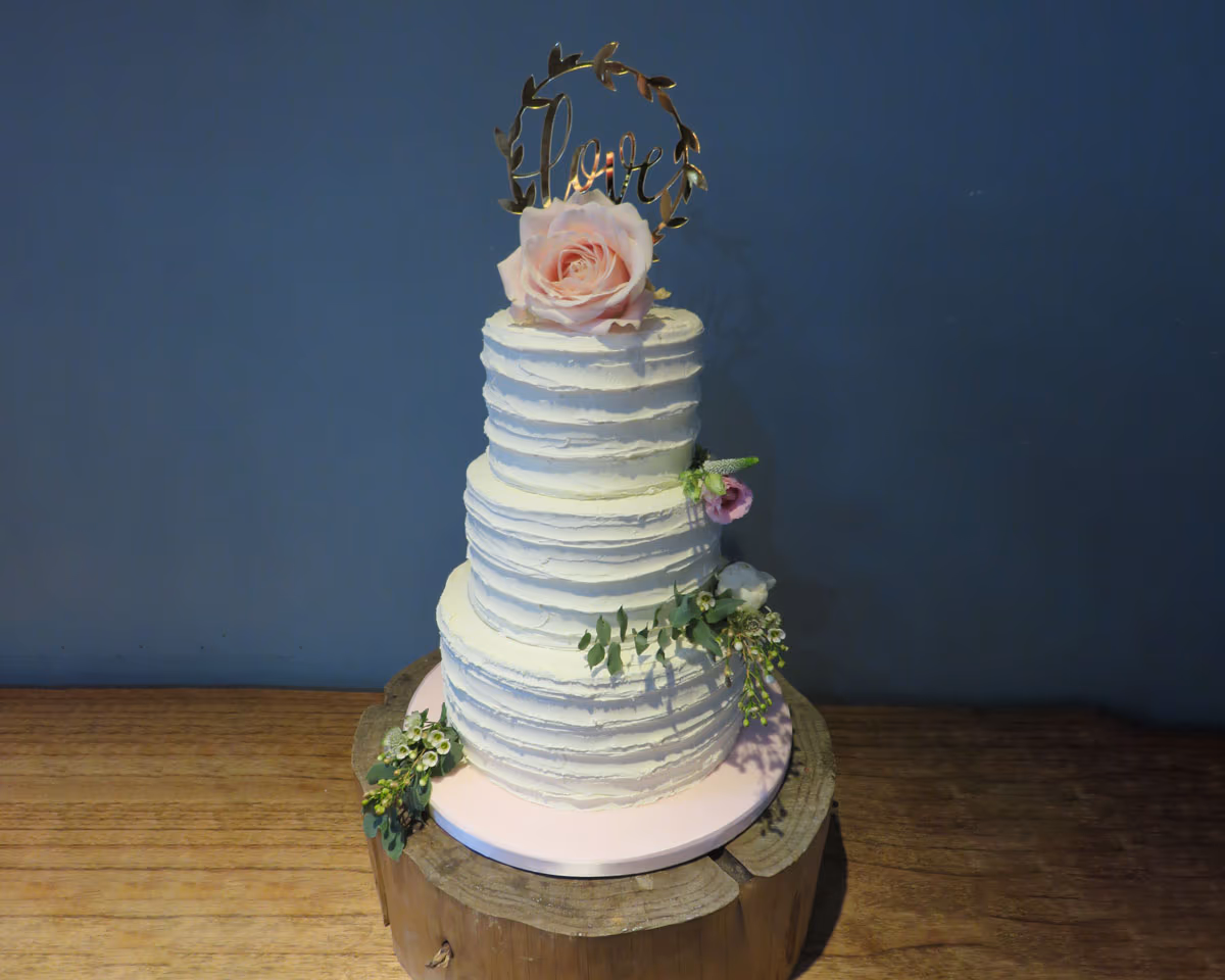 A three-tier wedding cake with white textured buttercream, adorned with a pink rose on top and a gold 'Love' cake topper, along with small floral accents and greenery on the sides. The cake sits on a pink board and a rustic wood log slice.