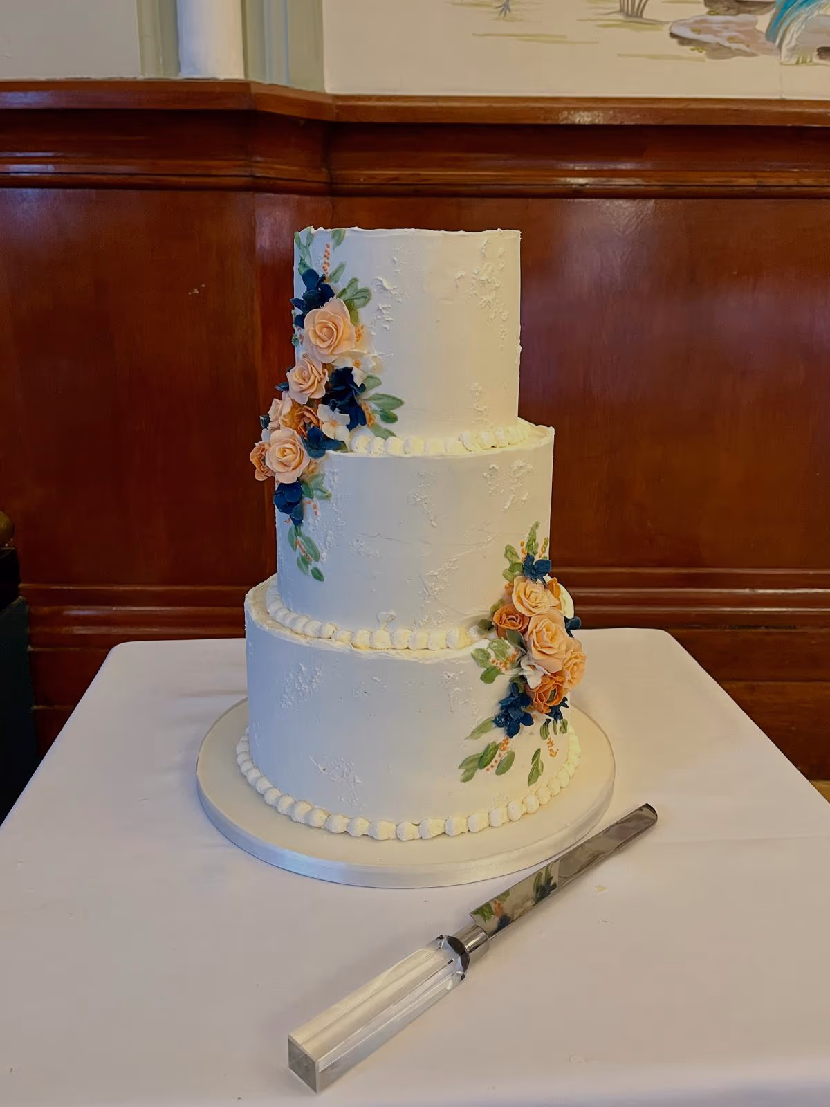 A three-tier white wedding cake with textured buttercream, decorated with cascades of peach and navy blue flowers and green leaves.