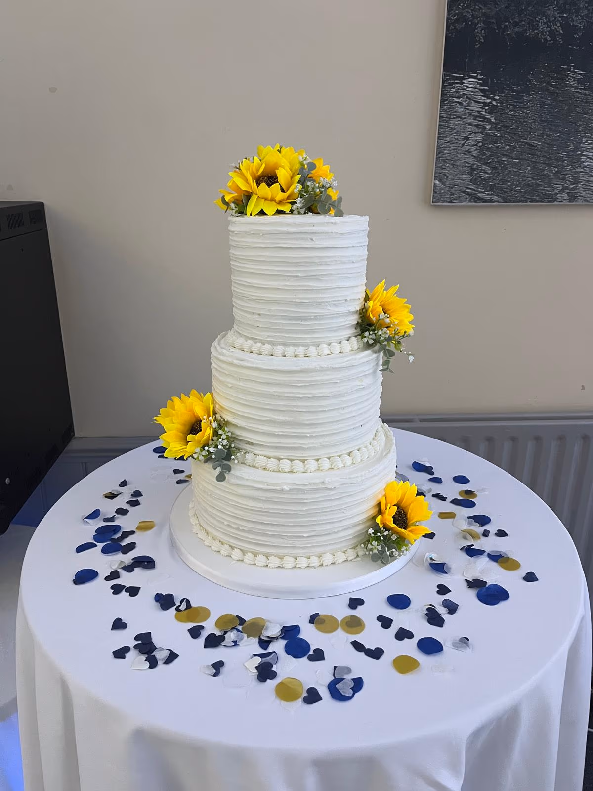 A three-tier wedding cake with white textured buttercream frosting, decorated with yellow sunflowers and greenery, placed on a white table with blue, gold, and heart-shaped confetti.