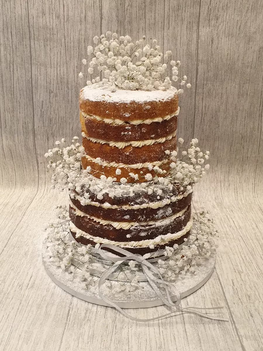 A two-tier naked wedding cake with visible cake layers and white cream filling, dusted with powdered sugar, and decorated with white baby's breath flowers and a silver ribbon.