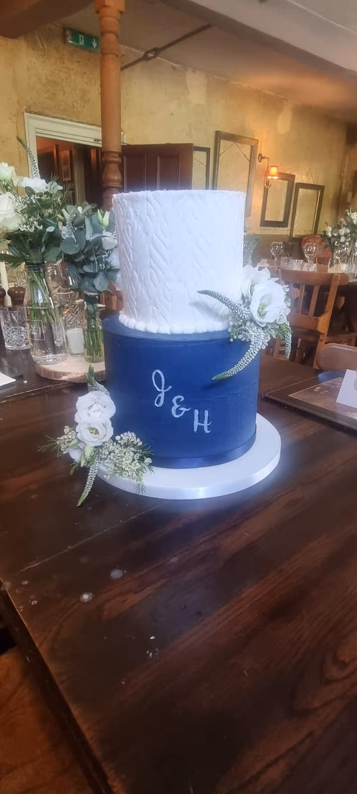 Two-tier wedding cake with a white textured top tier and a navy blue bottom tier featuring the initials J & H, decorated with white flowers and greenery, placed on a dark wooden table in a rustic venue.