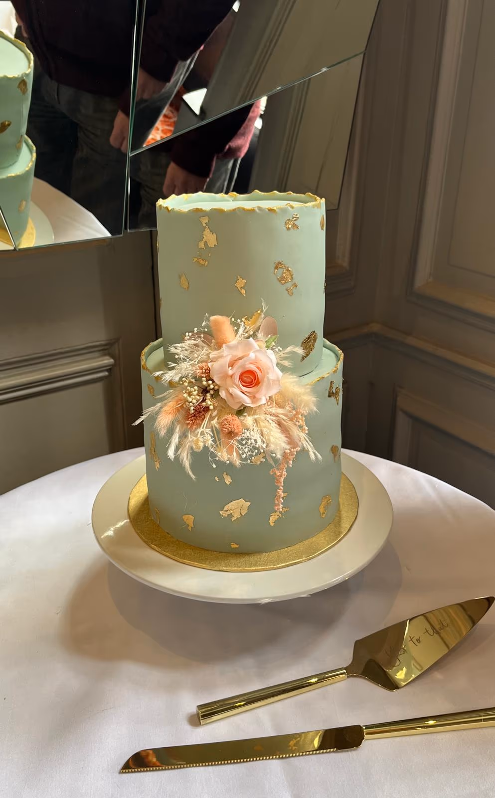A two-tier sage green wedding cake decorated with gold leaf and a floral arrangement featuring a pink rose, orange blossoms, and dried feathery grasses, displayed on a white table with gold cake serving utensils.