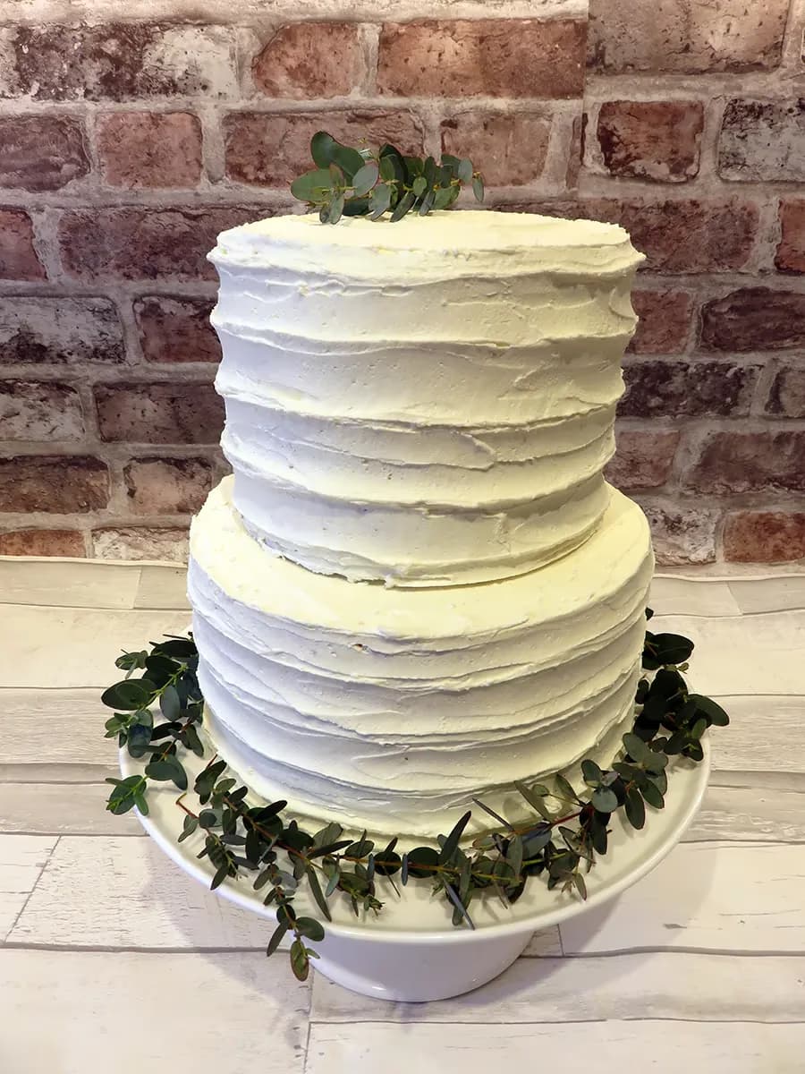 A two-tier white wedding cake with textured buttercream frosting, adorned with eucalyptus leaves on top and around the base, resting on a white cake stand.