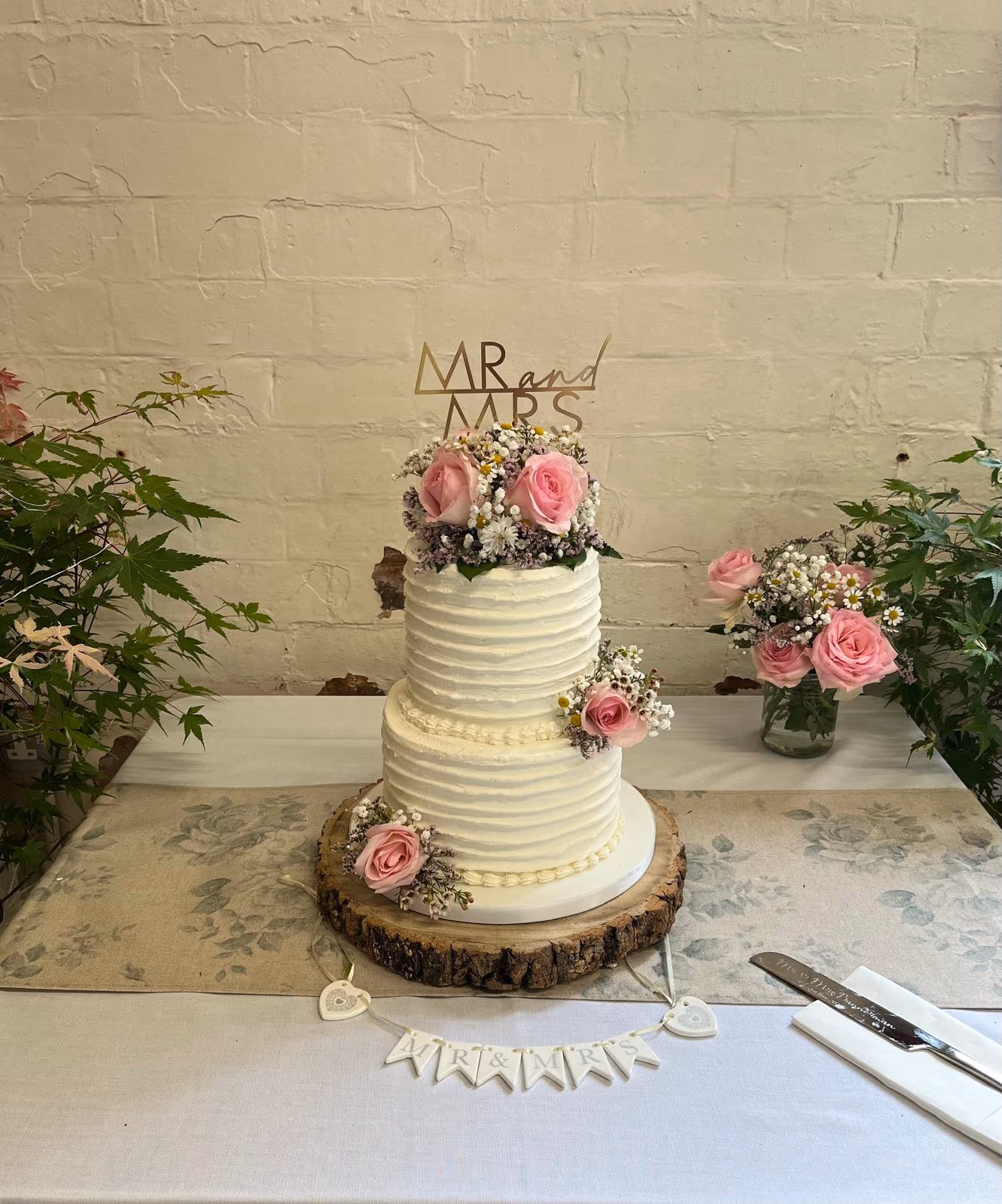 A two-tier wedding cake with textured white buttercream, decorated with pink roses and small white and purple flowers, topped with a gold 'MR and MRS' topper, sitting on a rustic wooden stand.