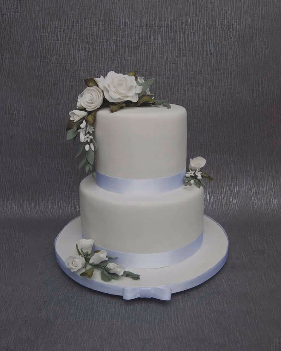 Two-tier white wedding cake decorated with light blue satin ribbons, white sugar roses, white berries, and green foliage cascading down the top tier and arranged at the base.