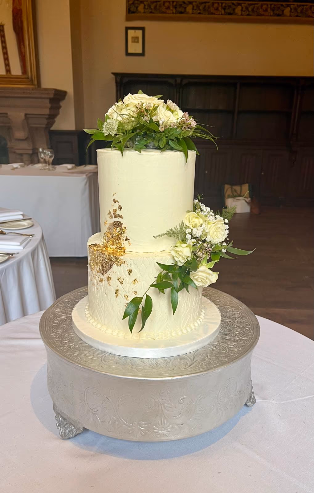 A two-tier wedding cake with smooth white cream frosting, featuring gold leaf accents cascading down the side of the bottom tier. The cake is decorated with bouquets of white roses and green foliage on top and on the side of the lower tier, which has a subtle textured pattern. The cake sits on an ornate silver cake stand.