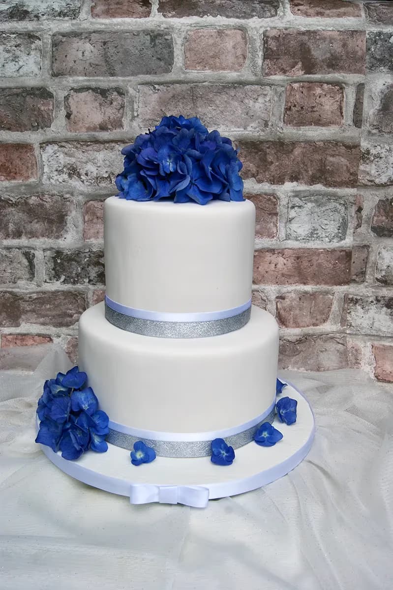 A two-tier white fondant wedding cake decorated with vibrant blue hydrangea flowers on top and cascaded on the base, accented with silver glitter ribbons around each tier.
