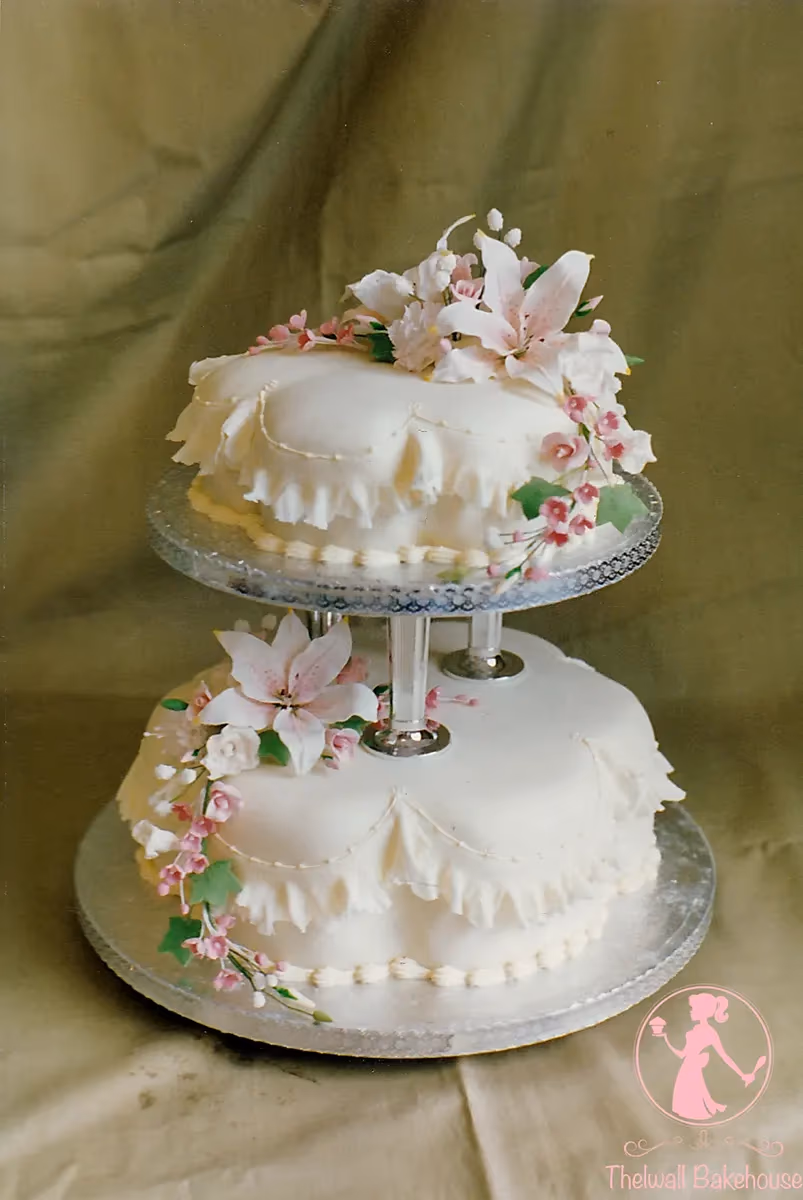 A white two-tier wedding cake with ruffled icing, decorated with pink and white lilies and small pink flowers with green leaves, displayed on a clear glass stand against a muted green fabric background.