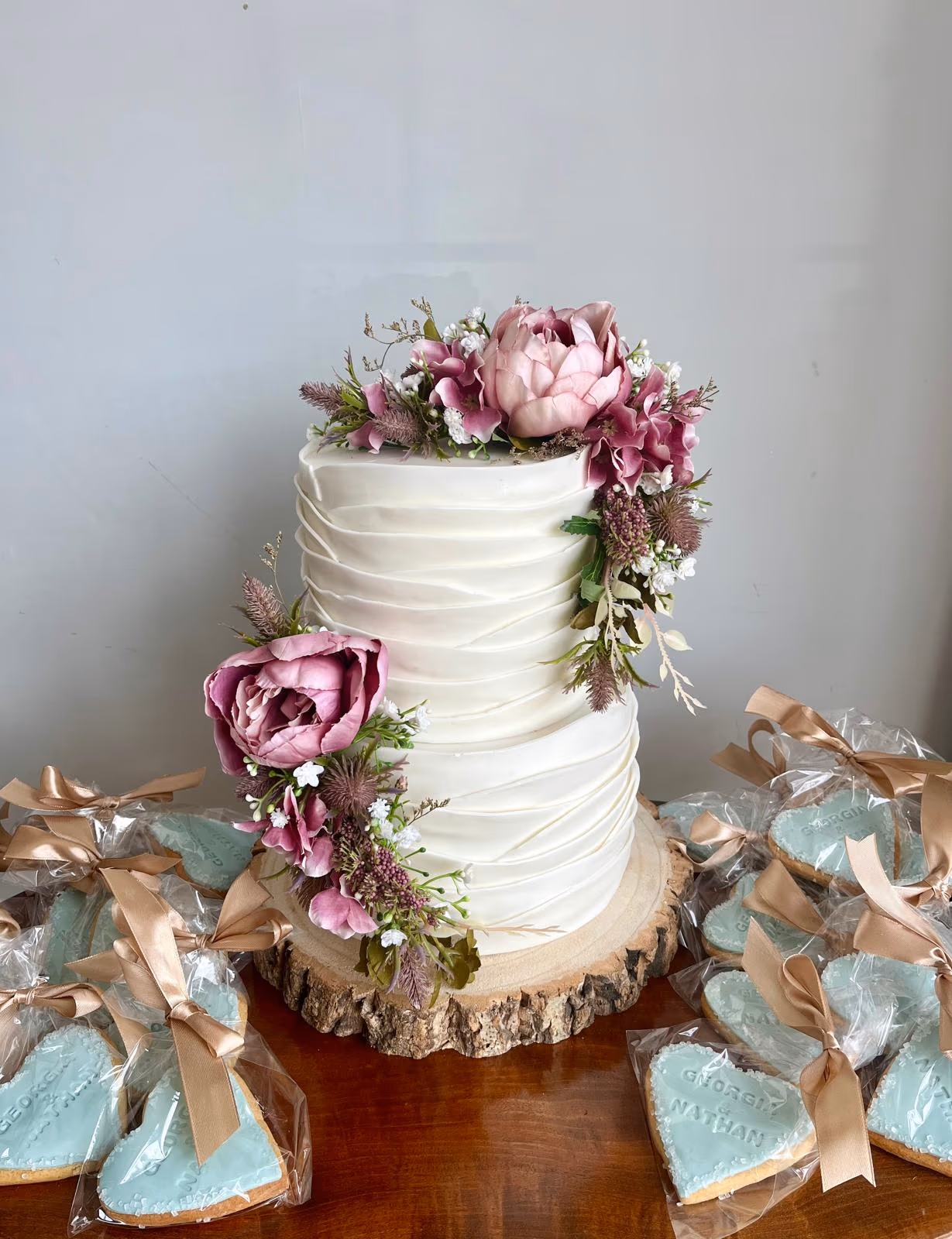 Two-tier white wedding cake with textured buttercream and pink and purple floral decorations, displayed on a wood slice stand, surrounded by heart-shaped light blue iced cookie wedding favors.