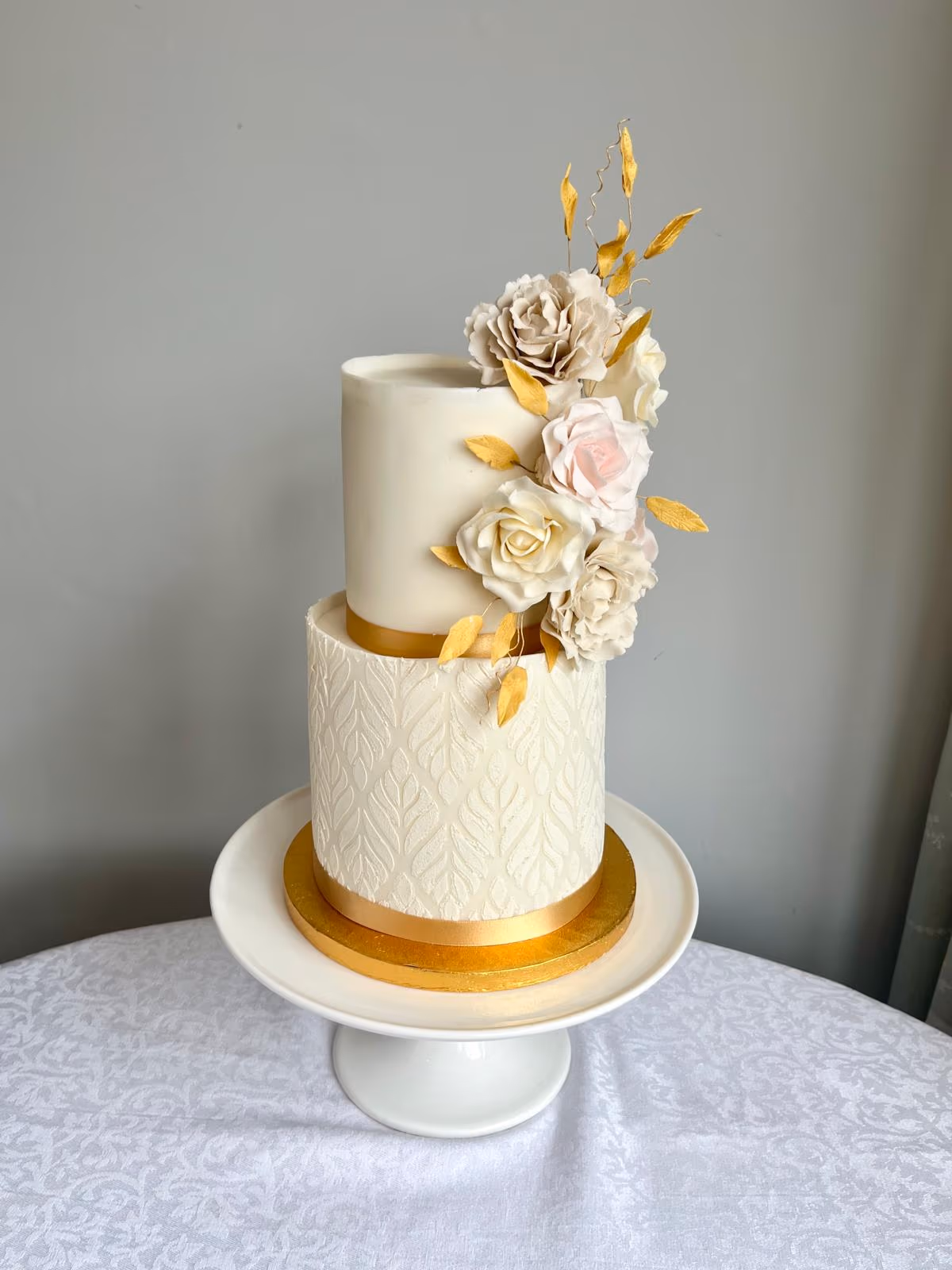 A two-tier white wedding cake featuring a textured leaf pattern on the bottom tier and a smooth top tier, each with a gold ribbon around its base. The cake is decorated with an arrangement of white, cream, and blush pink sugar roses and delicate golden leaves, presented on a white pedestal cake stand.