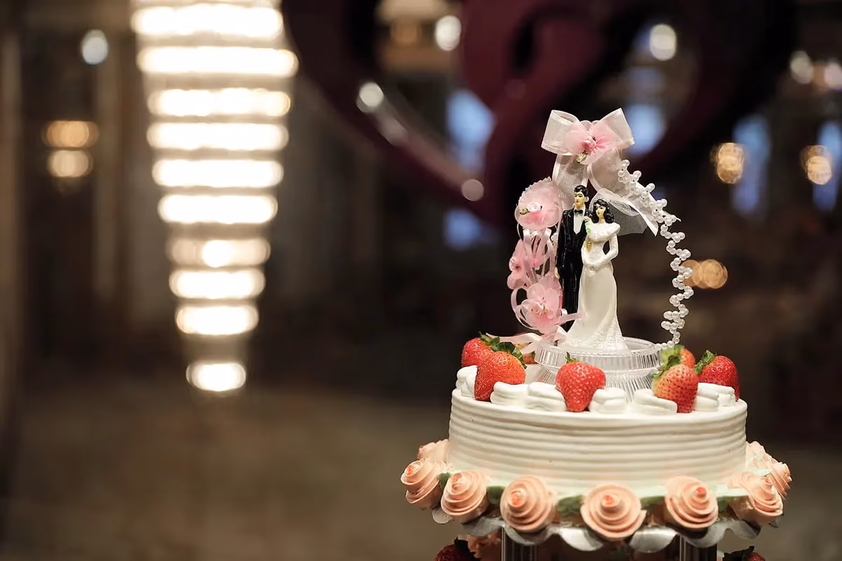 A white wedding cake with ribbed frosting, decorated with fresh strawberries and pink rose-shaped frosting, topped with a bride and groom figurine adorned with pink bows and a string of pearls.