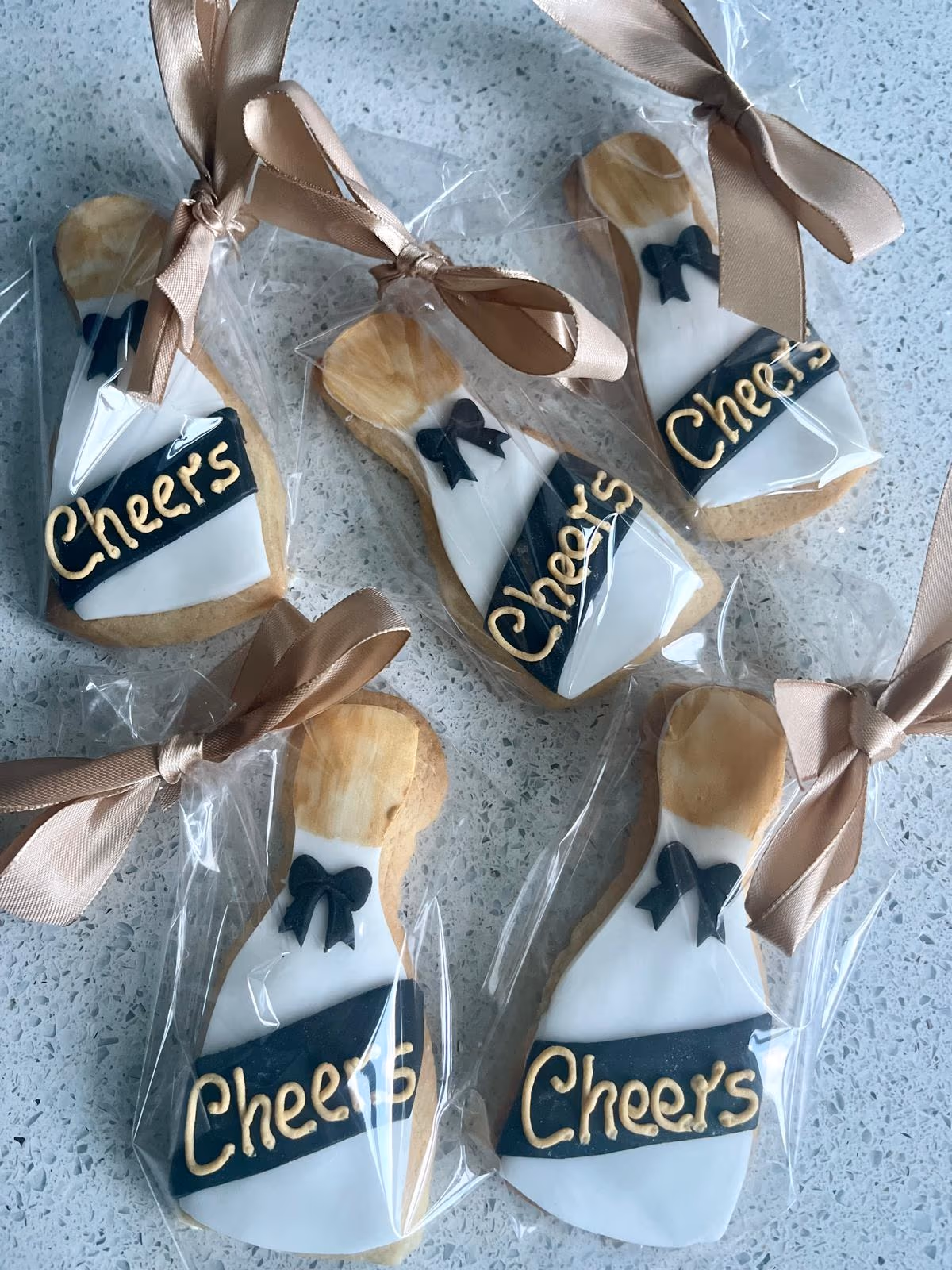 Five individually wrapped champagne bottle-shaped biscuits, decorated with white icing, a black label with gold 'Cheers' text, and a black bow, each tied with a gold ribbon.